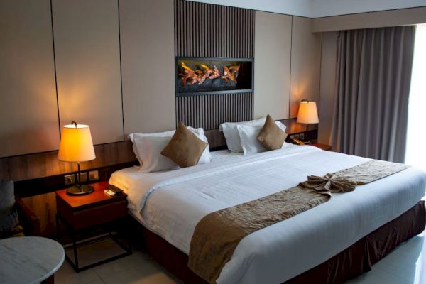 A well-lit hotel room with a large, neatly made bed, two bedside tables with lamps, a wall-mounted decor above the bed, and curtains.