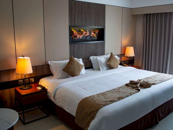 A well-lit hotel room with a large, neatly made bed, two bedside tables with lamps, a wall-mounted decor above the bed, and curtains.