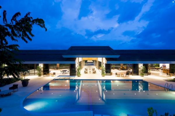 A luxurious property with modern architecture features a large, illuminated swimming pool and a spacious open-air seating area, viewed at twilight.
