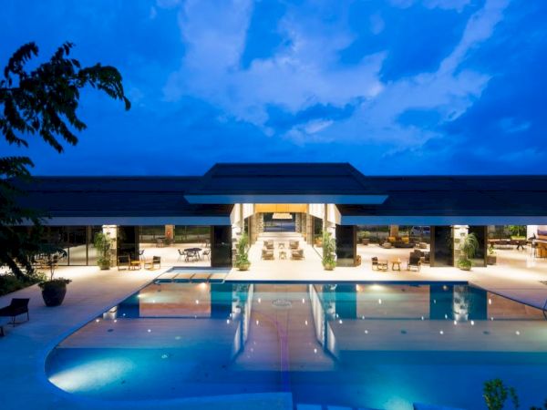 A luxurious property with modern architecture features a large, illuminated swimming pool and a spacious open-air seating area, viewed at twilight.