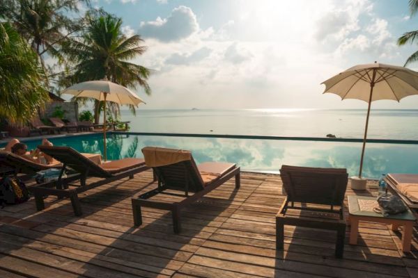 The image shows a picturesque tropical poolside with lounge chairs, parasols, palm trees, and a serene view of the sea under a partly cloudy sky.
