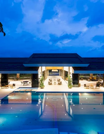 The image shows a luxurious, illuminated building with a large swimming pool in the foreground under a blue evening sky.