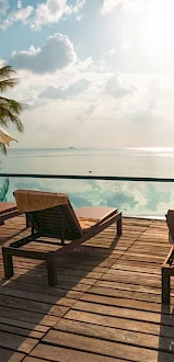 Lounge chairs and umbrellas by a swimming pool, overlooking a scenic ocean view with palm trees and a partially cloudy sky in the background.