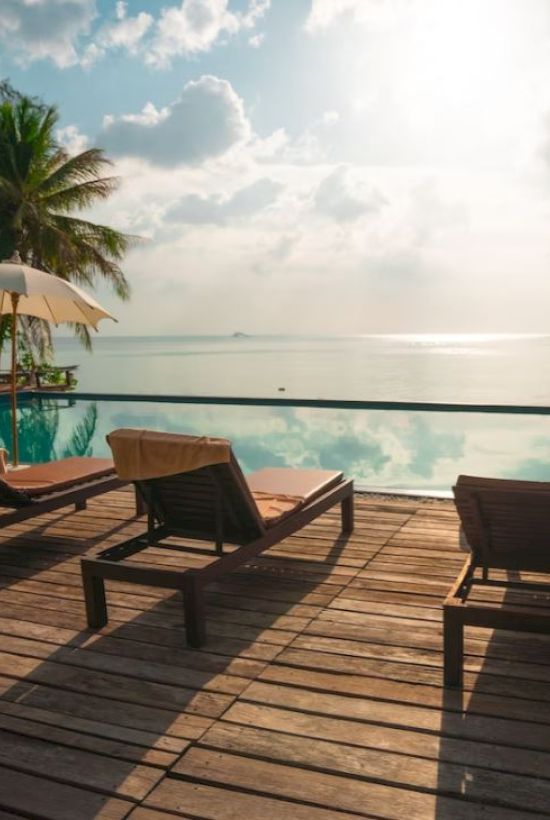 A serene poolside scene with lounge chairs, umbrellas, a wooden deck, and a view of the ocean under a partly cloudy sky.