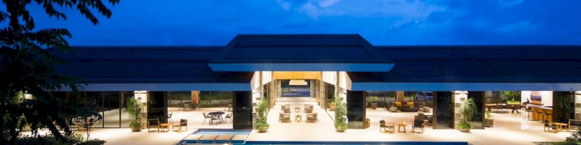 A luxurious, well-lit building with an expansive swimming pool in front, set against a dusk sky with scattered clouds.