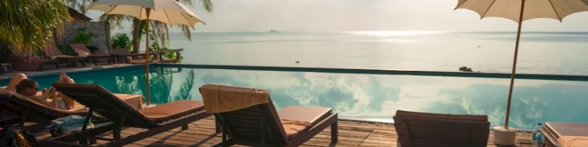Poolside scene with lounge chairs, umbrellas, and palm trees overlooking a serene body of water under a partly cloudy sky.