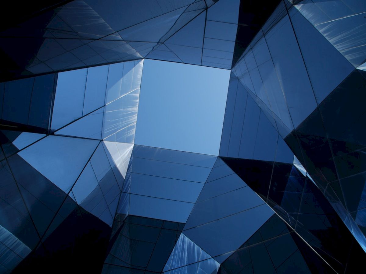 The image shows an upward view of a modern glass building with reflective surfaces creating geometric shapes against the blue sky.