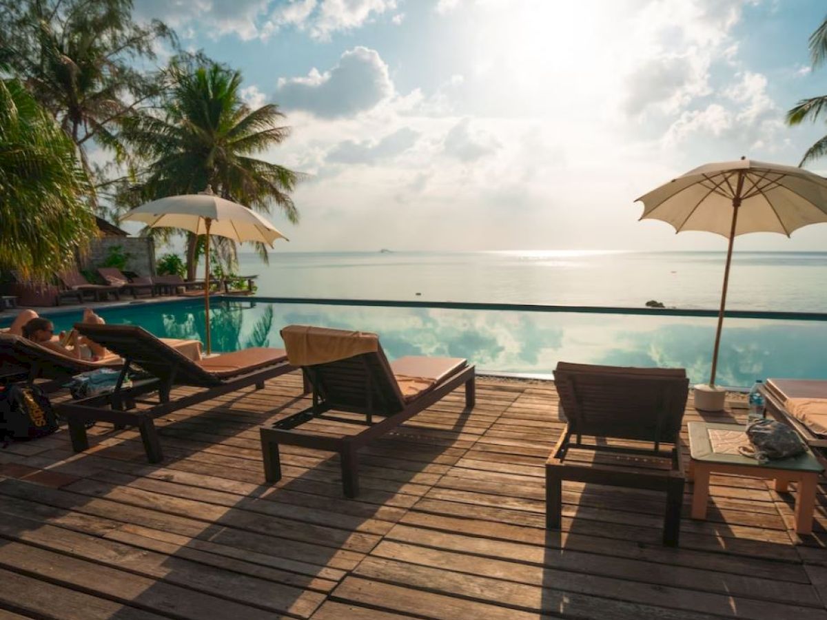 The image shows a serene poolside area with wooden loungers under umbrellas, overlooking a calm sea with a beautiful, partly cloudy sky.