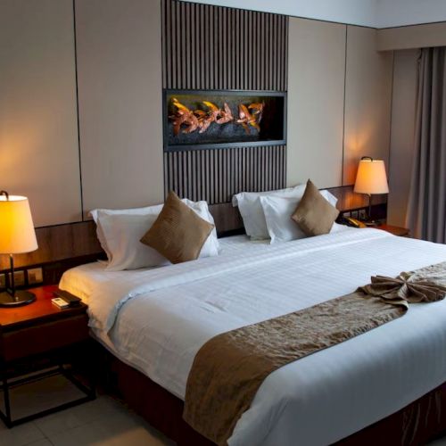 A neatly arranged hotel room with a large bed, two side tables with lamps, decorative pillows, and a wall-mounted art piece above the bed.