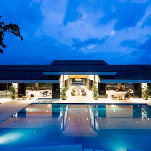 A well-lit modern building with large glass windows overlooks a serene, illuminated swimming pool against a twilight sky.