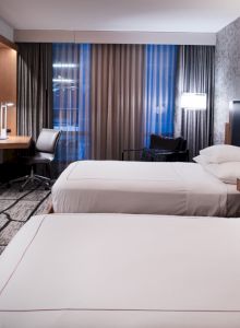 A modern hotel room with two beds, a flat-screen TV, a work desk with a chair, and floor-to-ceiling windows covered with curtains.