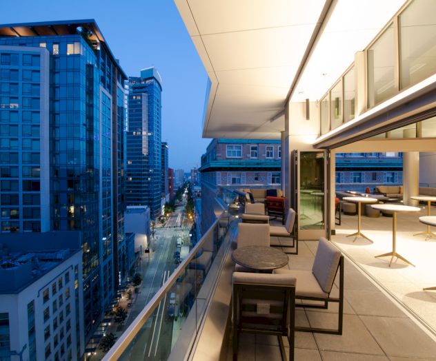 A modern balcony with a view of a cityscape at dusk, featuring outdoor seating and tables, with buildings and lit streets in the background.