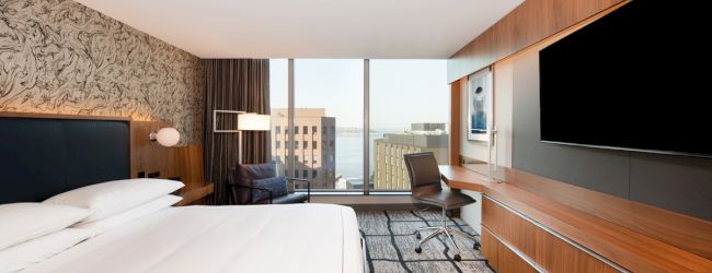 A modern hotel room with a large bed, flat-screen TV, desk, and chair, featuring a window with a cityscape view and contemporary decor.