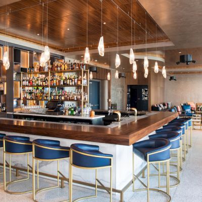 A modern bar with a wooden counter, blue bar stools, and an extensive selection of liquor bottles. Pendant lights hang from the ceiling.