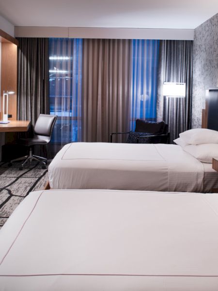 A modern hotel room with two double beds, a wall-mounted TV, a workspace with a chair, bedside lamps, and large windows with curtains.