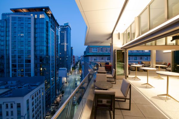 A modern balcony overlooking a cityscape at dusk, with tables and chairs arranged for seating, and city lights illuminating the streets below.