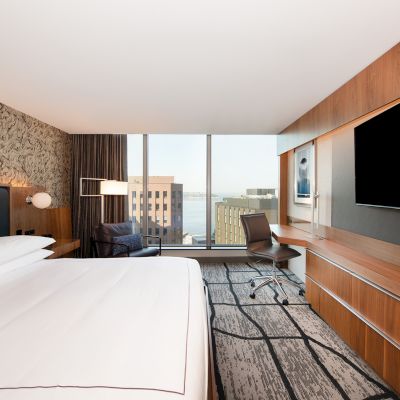 A modern hotel room features a large bed, wall-mounted TV, desk with chair, floor-to-ceiling window, and cityscape view beyond.
