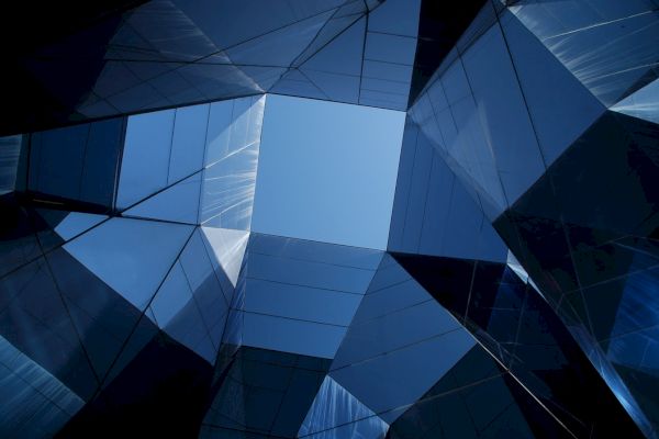 The image shows a geometric perspective looking up at a sky through multiple reflective glass surfaces of a modern building.