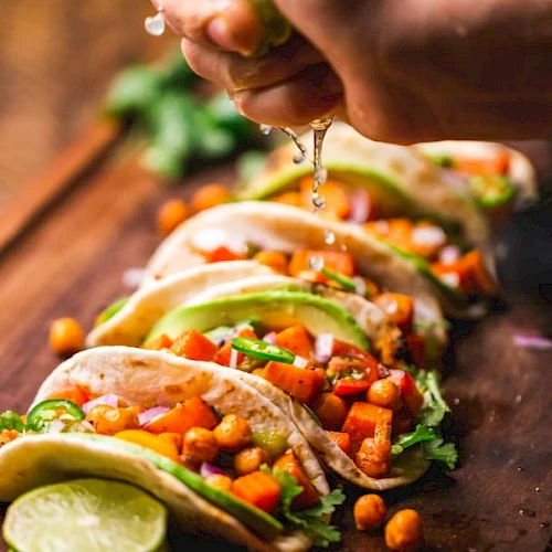 A hand is squeezing a lime over a row of delicious-looking vegetarian tacos filled with chickpeas, avocado, and other fresh ingredients.