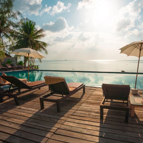 The image shows a serene poolside with lounging chairs, umbrellas, and a beautiful view of the sea under a partly cloudy sky.