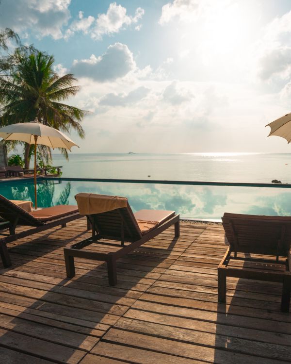 The image shows a serene poolside with lounging chairs, umbrellas, and a beautiful view of the sea under a partly cloudy sky.
