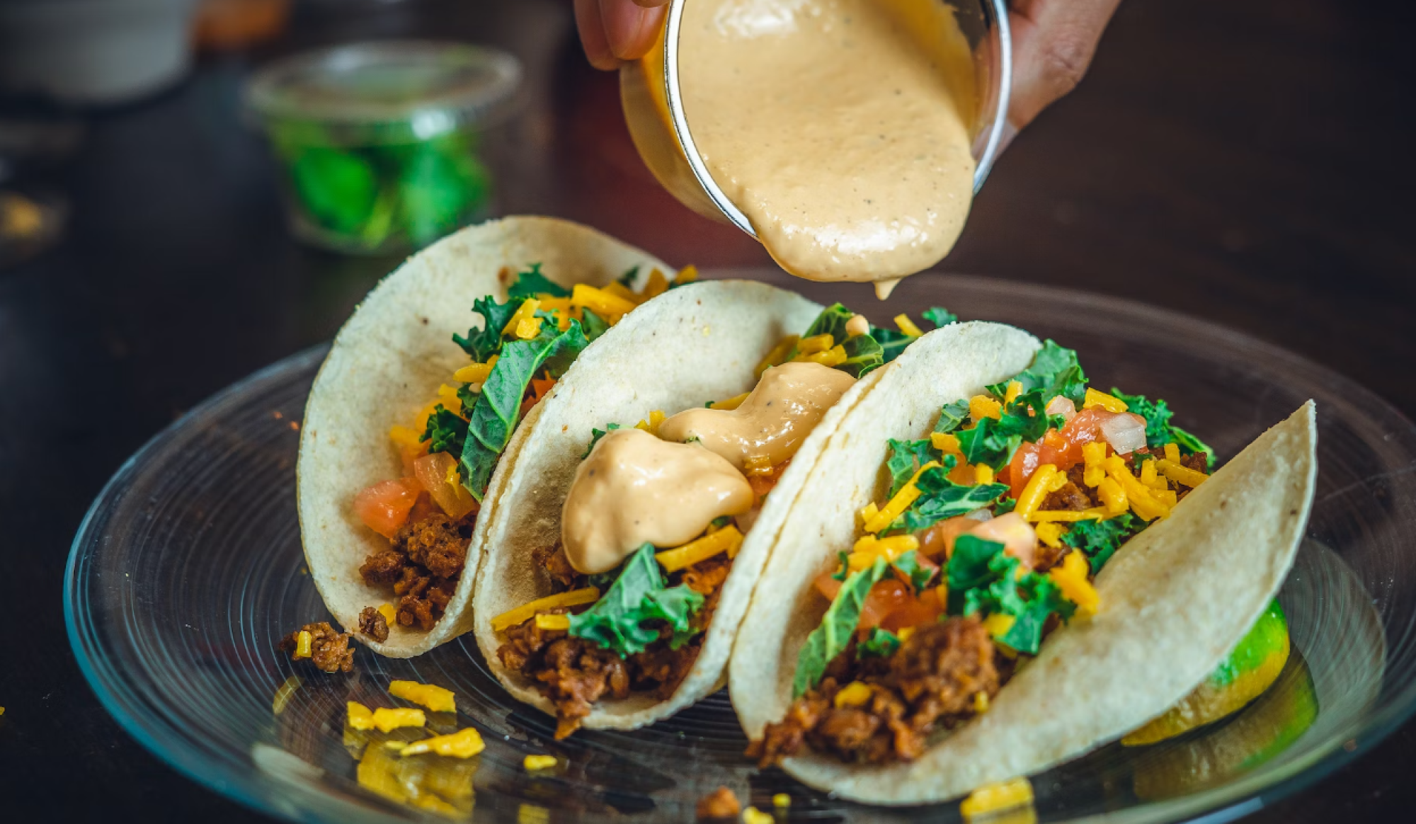 Three tacos filled with ground beef, lettuce, tomato, and shredded cheese are being topped with a creamy sauce from a small cup.