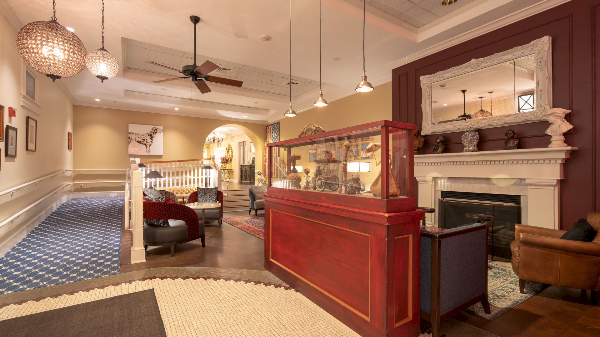 A warmly lit hotel lobby features a reception desk, cozy seating areas, a large mirror above a fireplace, and patterned carpets throughout the space.