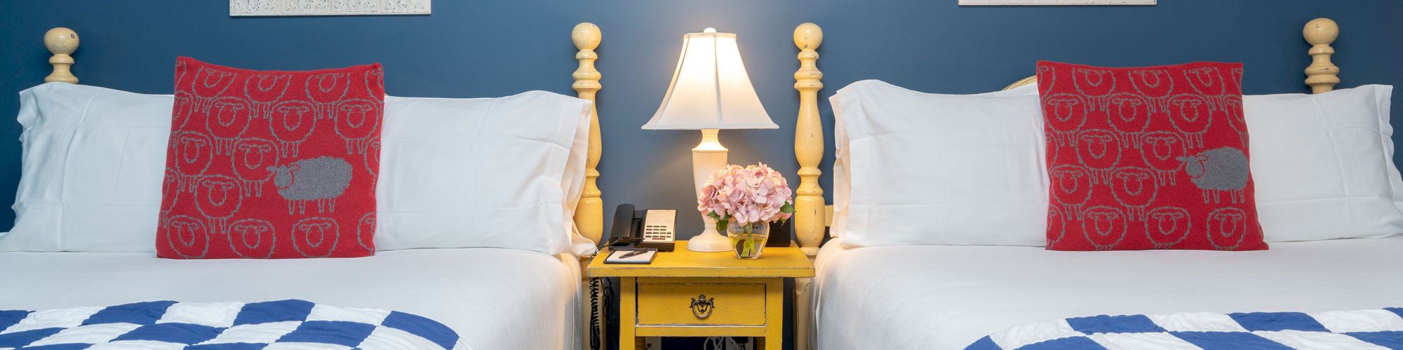 This image shows a cozy bedroom with two single beds, a yellow nightstand in the middle, and a lamp. The walls are blue with white square mirrors.