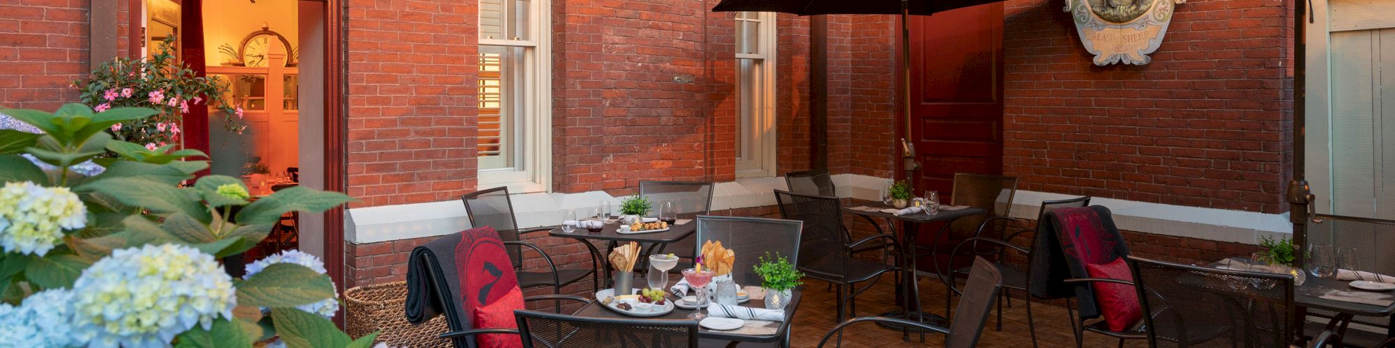 Outdoor dining area with string lights, tables with food, flowers, and umbrellas against a brick building with windows and plants.