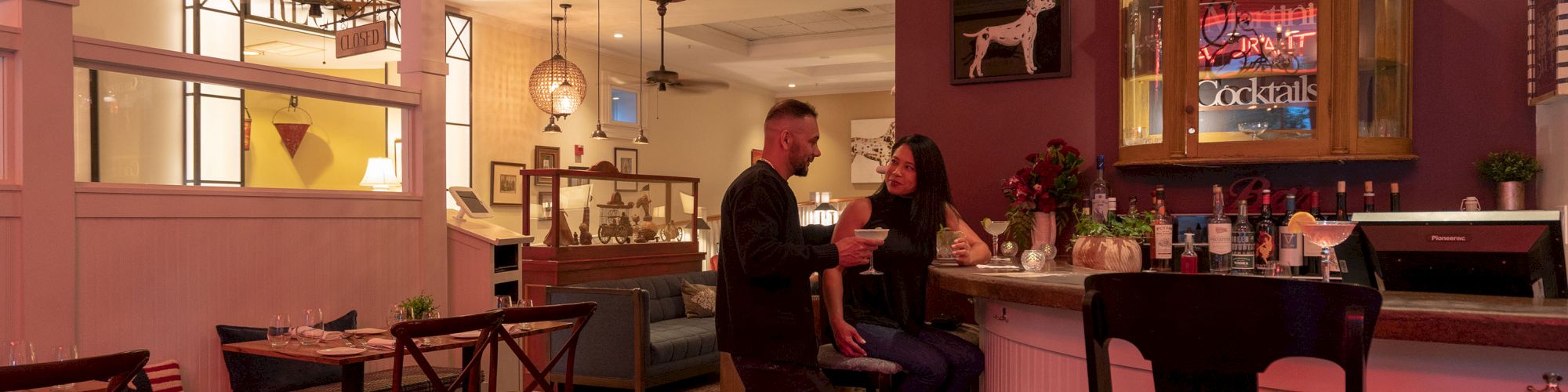 A couple is conversing at a bar in a cozy restaurant with wooden furniture and warm lighting. The bar shelves display various bottles and decor.