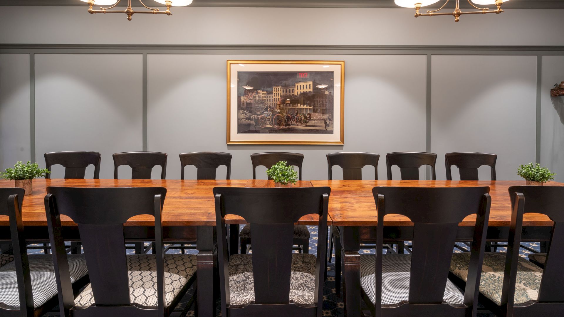 A long table with multiple chairs, indoor lighting, small plants, and a framed wall picture in what appears to be a meeting room.