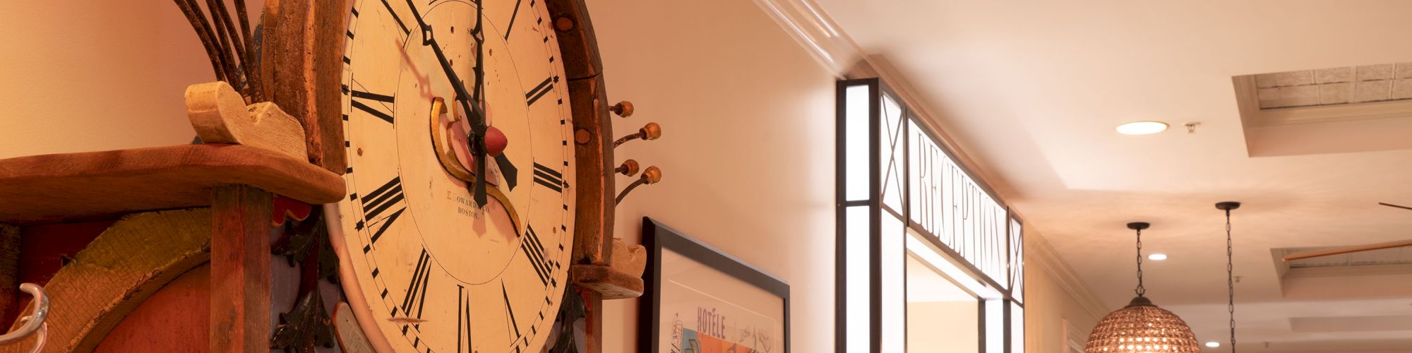 An ornate clock hangs on the left, with art pieces and a brightly lit hallway on the right, featuring pendant lights and framed art.