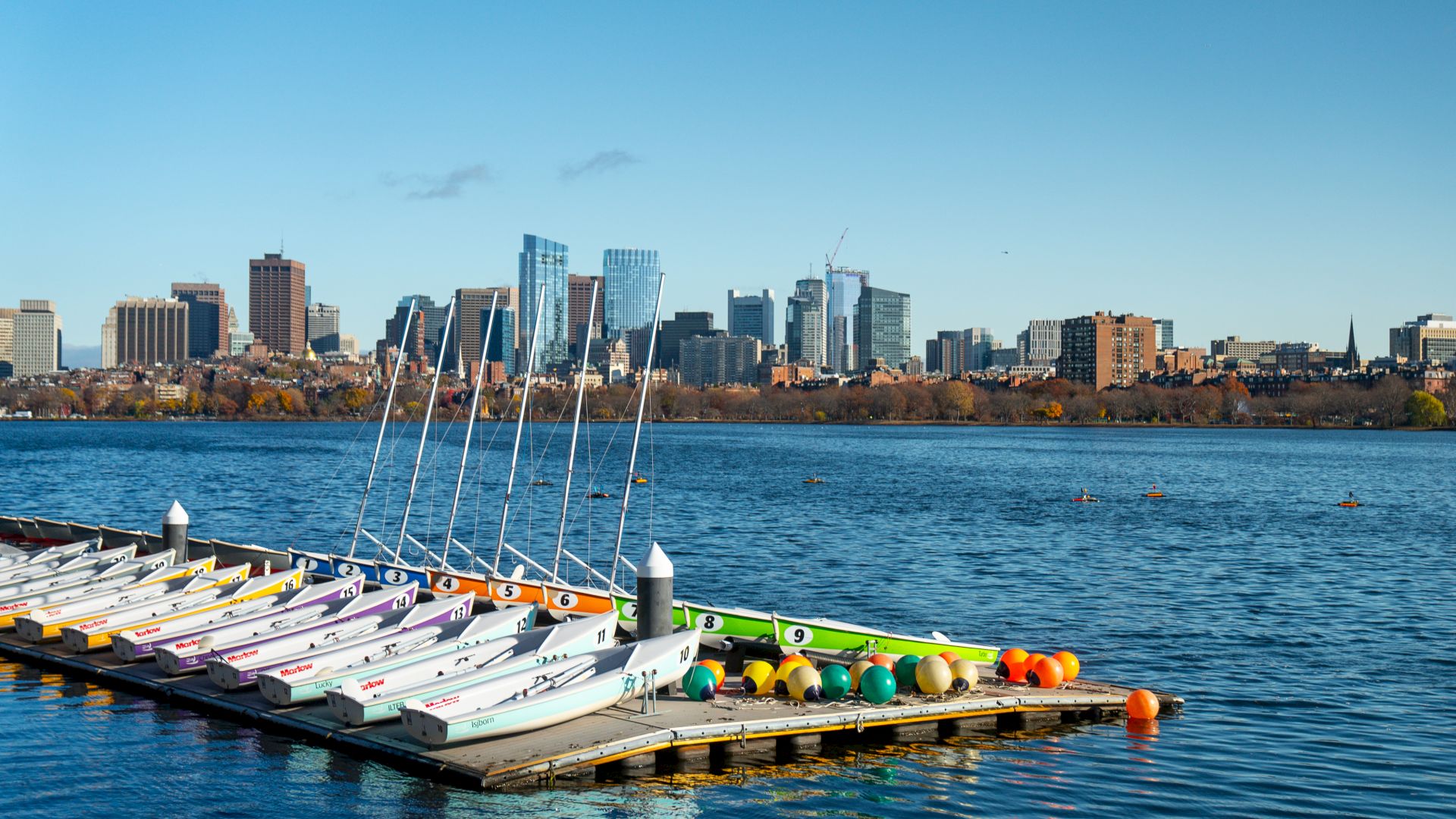A dock with sailboats is in the foreground, overlooking a river and city skyline under a clear blue sky.