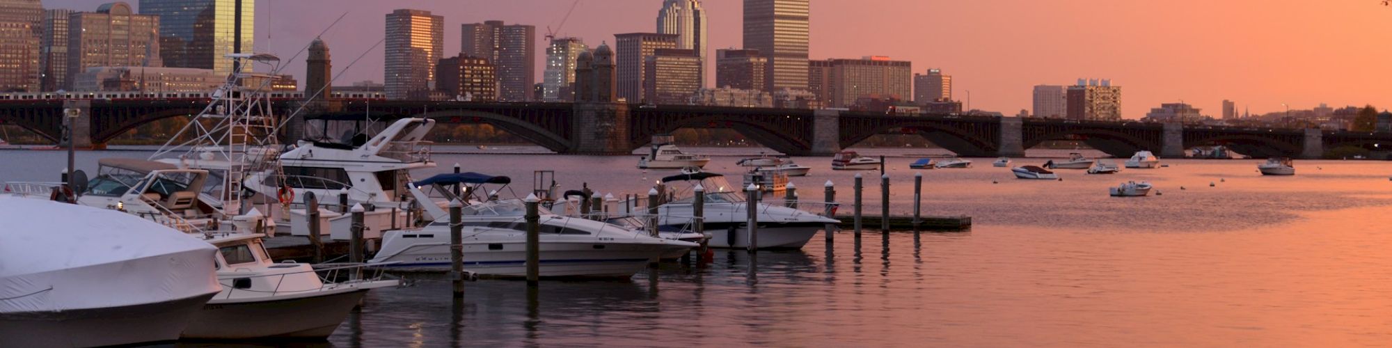 A sunset view of a city's waterfront with multiple boats docked, skyscrapers in the background, and a vibrant orange-pink sky reflecting in the water.