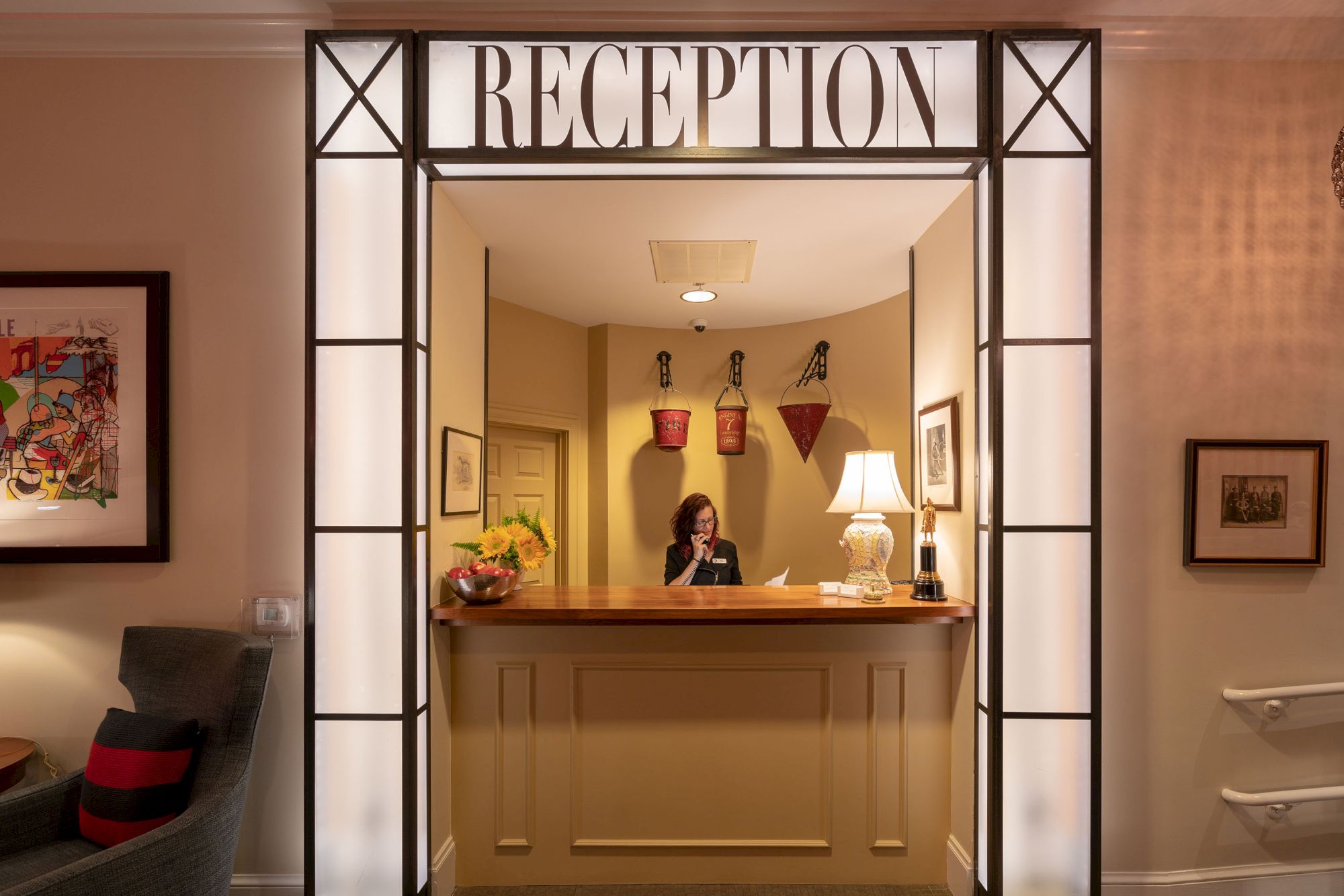 A reception area with a receptionist at a desk, decorated with wall art, a table lamp, and a bowl of fruit. The space is well-lit and welcoming.