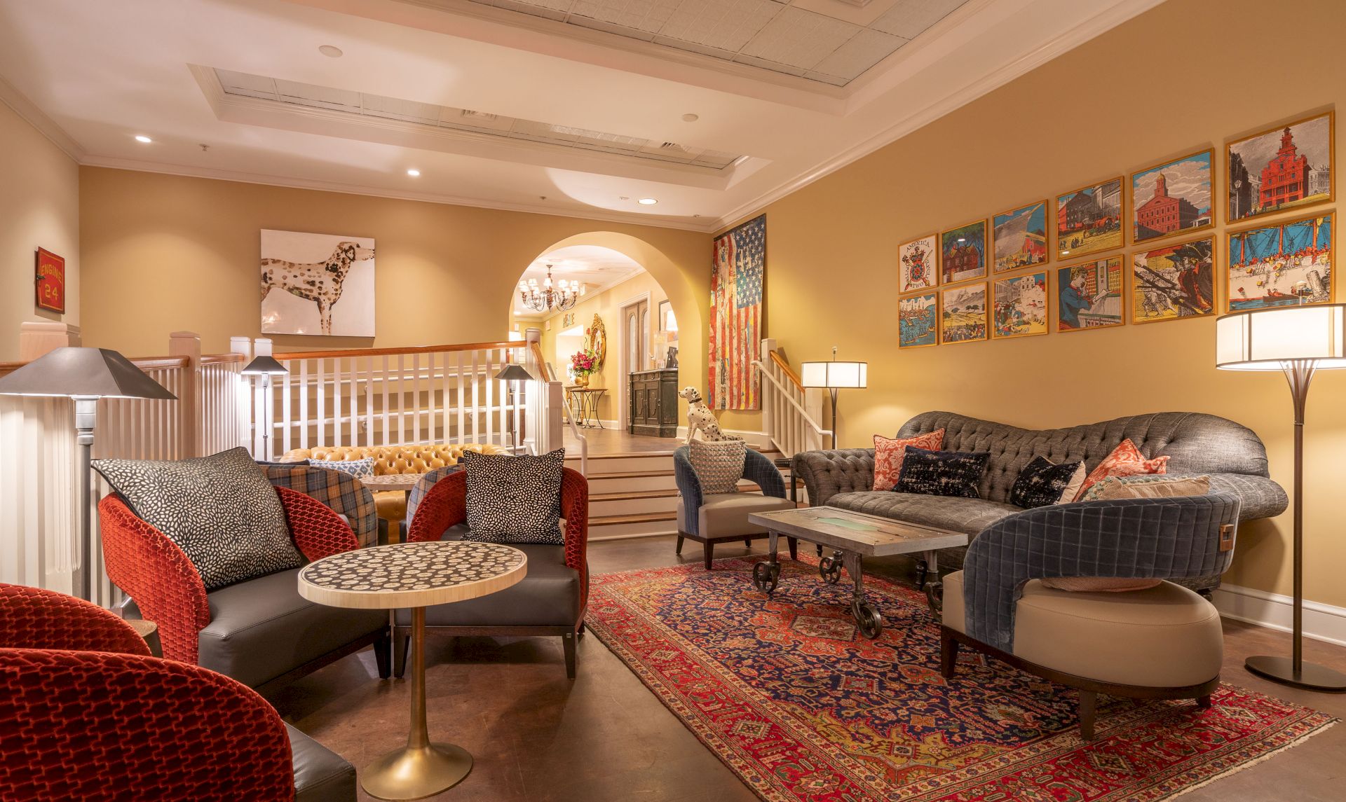 The image shows a cozy, well-lit lounge area with comfortable seating, colorful artwork on the walls, and a patterned rug.