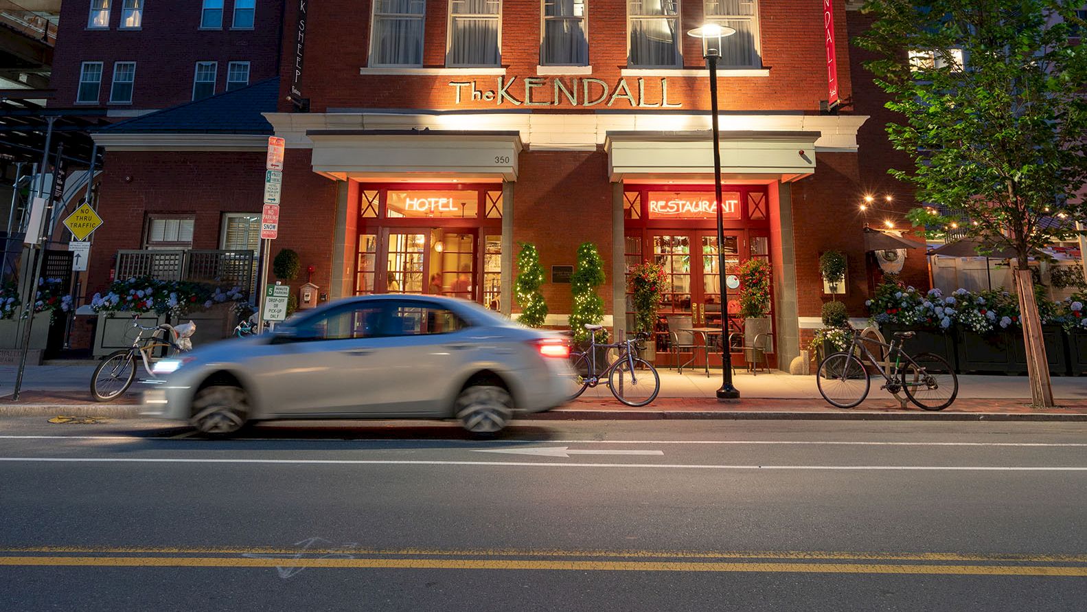 The image shows a brick building with a tower, lit at night. A car is passing in front, and there are bicycles parked by the building.