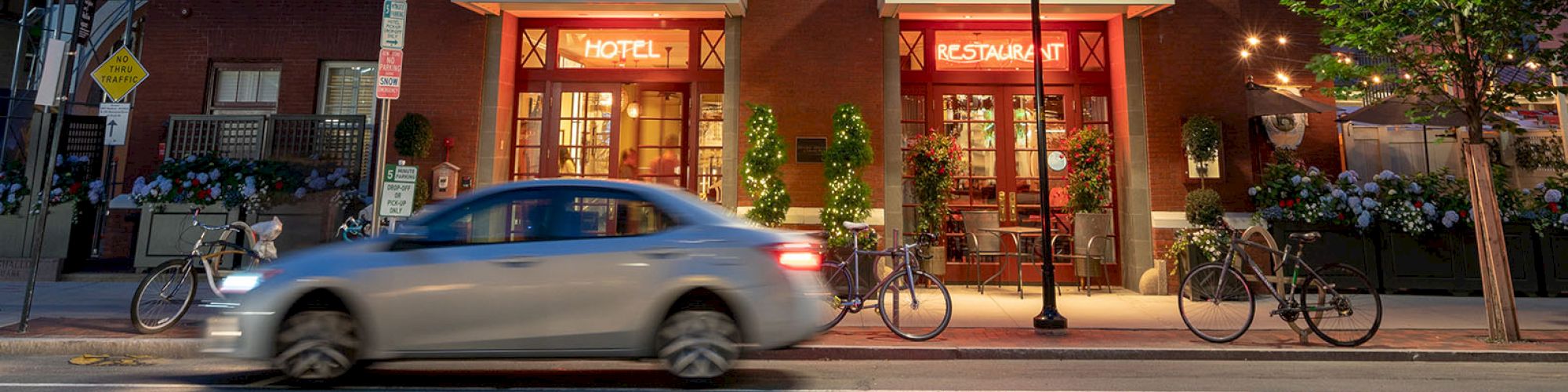 The image shows a brick building with a tower, lit at night. A car is passing in front, and there are bicycles parked by the building.