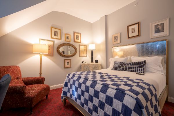 A cozy bedroom with a checkered bedspread, red armchair, multiple framed pictures on the walls, and two table lamps providing warm lighting.