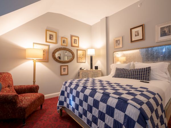 A cozy bedroom with a checkered bedspread, red armchair, multiple framed pictures on the walls, and two table lamps providing warm lighting.