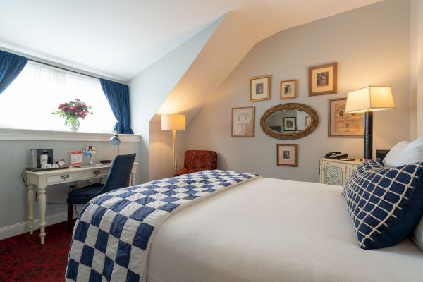 A cozy bedroom with a bed, blue and white quilt, a desk, chair, wall art, two lamps, a window with blue curtains, and a floral arrangement.