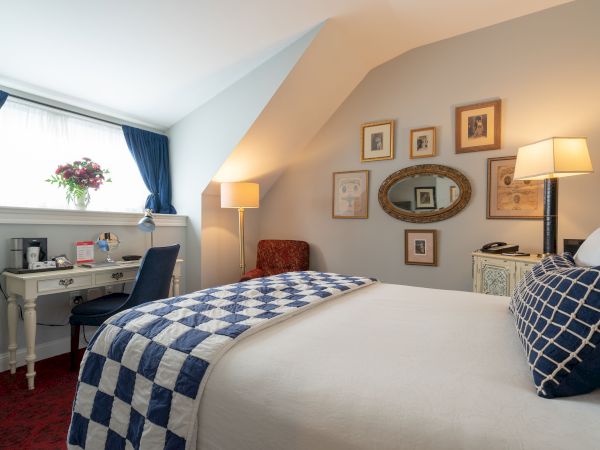 A cozy bedroom with a bed, blue and white quilt, a desk, chair, wall art, two lamps, a window with blue curtains, and a floral arrangement.