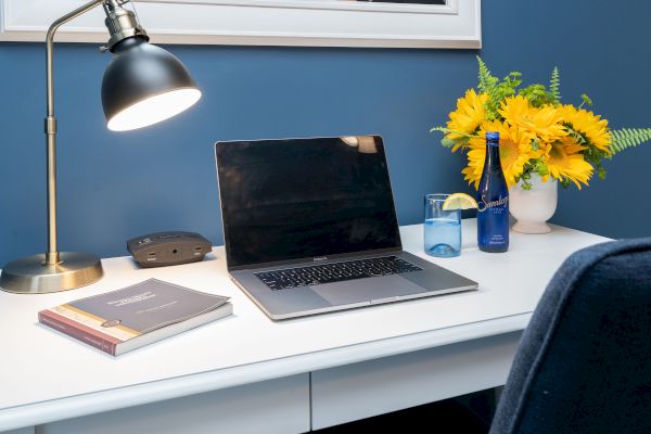 A tidy desk setup with a laptop, lamp, books, and a drink beside a vase of yellow flowers against a blue wall, shot in a cozy workspace.