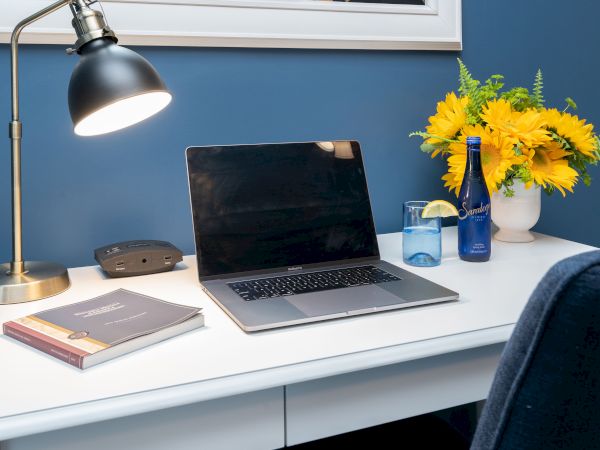 A tidy desk setup with a laptop, lamp, books, and a drink beside a vase of yellow flowers against a blue wall, shot in a cozy workspace.