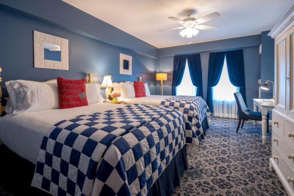 A cozy hotel room with two double beds, checkered blue and white bedspreads, red pillows, a ceiling fan, dark curtains, and a small desk area.