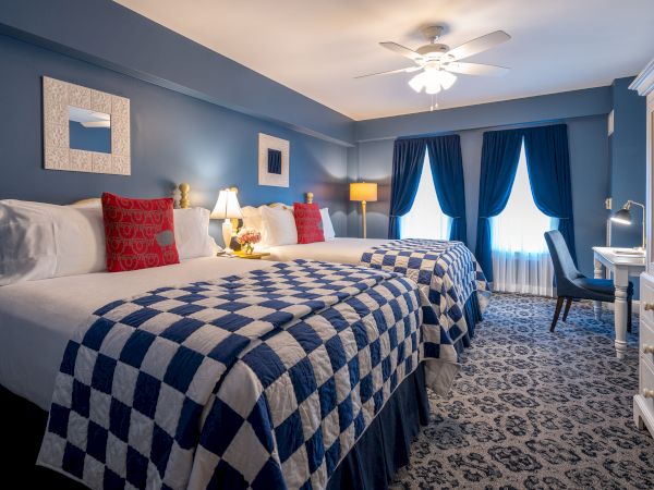 A cozy hotel room with two double beds, checkered blue and white bedspreads, red pillows, a ceiling fan, dark curtains, and a small desk area.