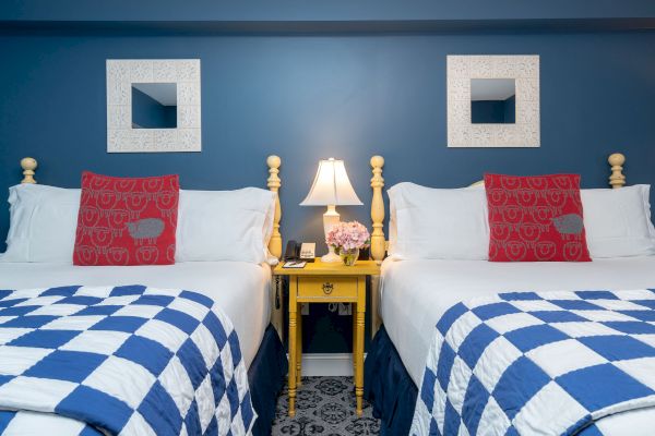 The image shows two neatly made beds with blue and white checkered bedding, red pillows, a yellow nightstand with a lamp, flowers, and two wall mirrors.