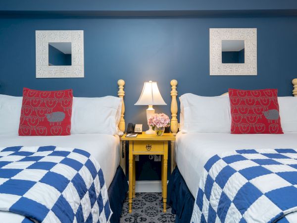 The image shows two neatly made beds with blue and white checkered bedding, red pillows, a yellow nightstand with a lamp, flowers, and two wall mirrors.