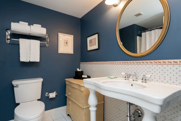 A bathroom with blue walls features a toilet, wall rack with towels, a mirror above a sink, and decorated with framed artwork and wall tiles.