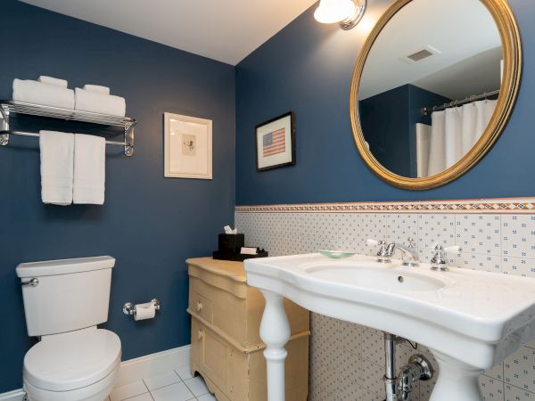 A bathroom with blue walls features a toilet, wall rack with towels, a mirror above a sink, and decorated with framed artwork and wall tiles.
